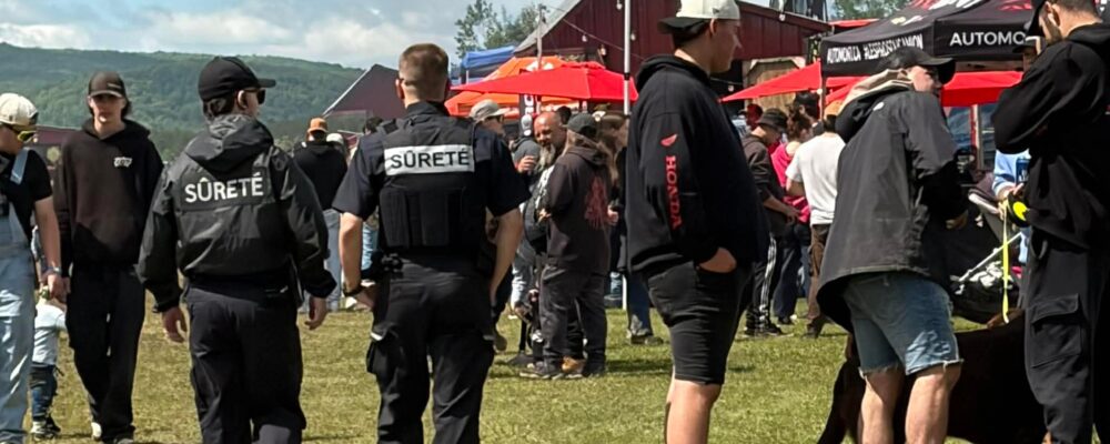 Security personnel in black jackets labeled 'Sûreté' stand among a crowd at an outdoor event with red tents and booths in the background.