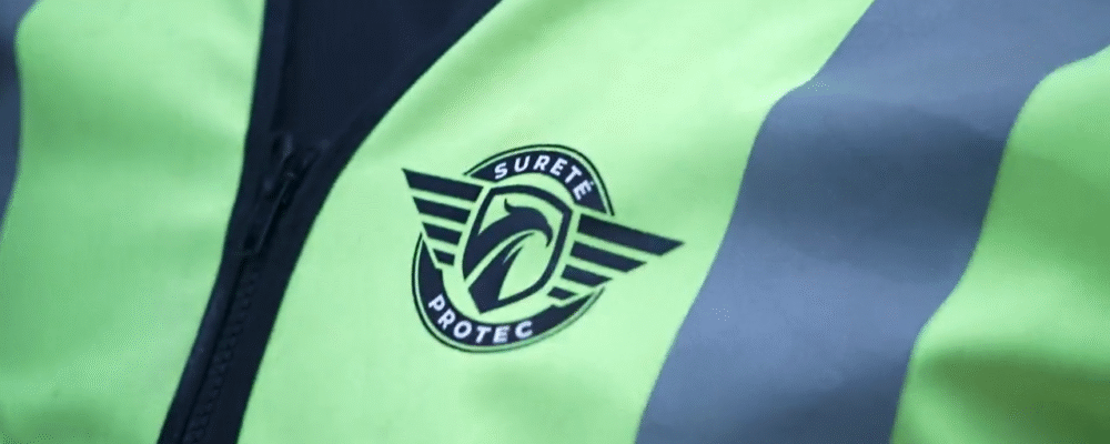 Close-up of a neon-yellow safety vest with a black circular badge reading 'Sureté Protec' and winged stripes beside the zipper.