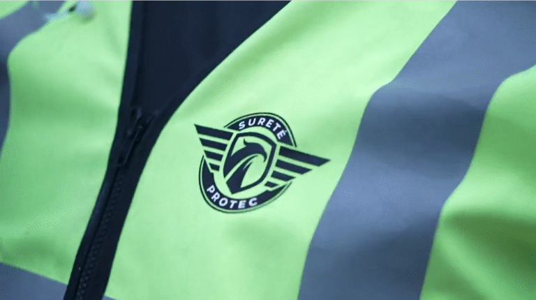 Close-up of a neon-yellow safety vest with a black circular badge reading 'Sureté Protec' and winged stripes beside the zipper.